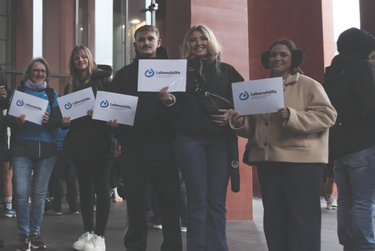 Mehrere Personen stehen nebeneinander. Jede Person hält ein Blatt Papier, auf dem das WMB-Logo abgedruckt ist.