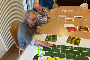 Zwei Bewohner sitzen an einem Tisch. Vor ihnen steht eine Pyramide mit verschiedenen Lebensmitteln, die sie sortieren oder zuordnen.