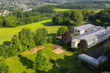 Luftaufnahme des Gebäudes des Hotel FIT mit Spielplatz, umgeben von Wald, Feldern und einer Ortschaft im Hintergrund