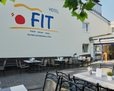 Außenansicht des Hotels FIT mit Logo sowie Restaurant-Außenbereich mit Tischen und Sitzgelegenheiten.